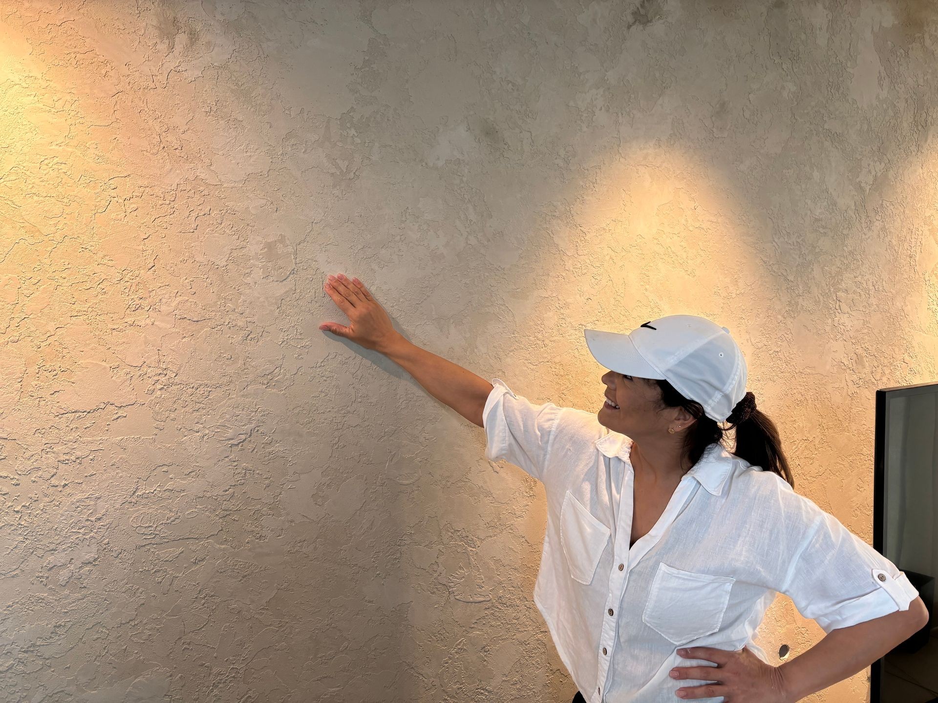 Person wearing a white shirt and a white cap, touching a textured beige wall under warm lighting.